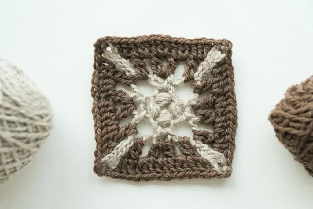 A handmade Compass Star Granny Square crocheted in two contrasting colors, featuring a raised four-puff center and four long pointed chain arms arranged like a compass rose inside a neat square border, photographed directly from above on a soft neutral background with gentle natural daylight, crisp