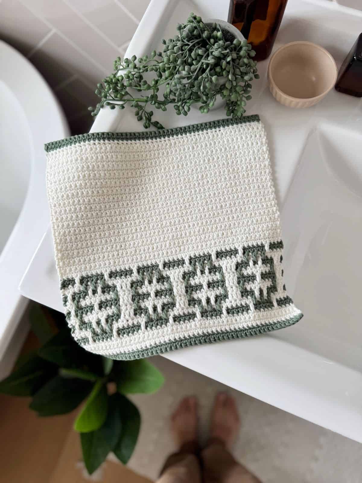 A handmade square inset mosaic crochet washcloth in soft cream and muted green worsted-weight yarn, featuring a clean geometric mosaic panel and solid single-crochet sections, photographed flat from above on a bright neutral surface with soft natural daylight, crisp stitch definition, balanced color